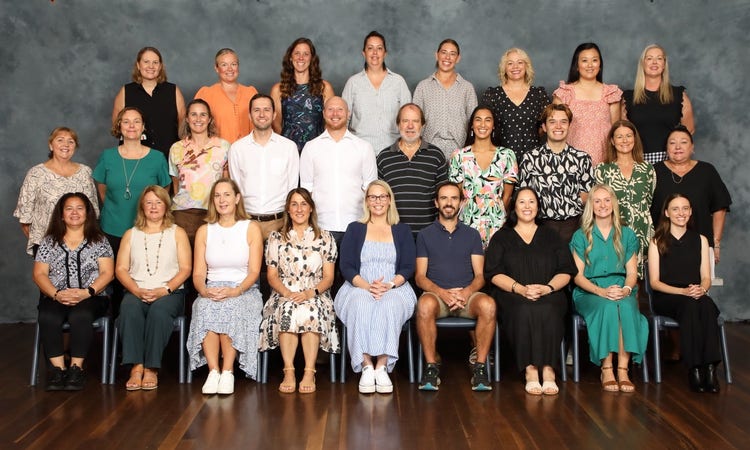 A formal group portrait of the Gosford East Public School staff and teachers shows three rows of adults seated and standing on polished wooden floors against a neutral studio backdrop. The staff are dressed in a range of professional and semi-casual outfits, including shirts, dresses, blouses, and slacks, presenting a unified team. This photograph represents the educators and support staff who make up the school community.