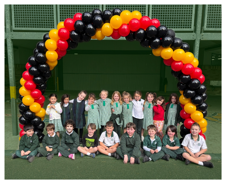A group of young children in green school uniforms sit and stand in two rows beneath a large balloon arch made of black, yellow, and red balloons inside a school playground.