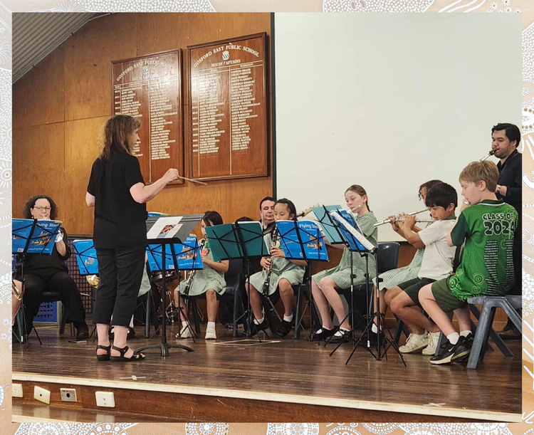 Students in green checkered uniforms and a teacher participate in a music ensemble on stage, playing wind instruments such as flutes and clarinets. The conductor guides the group as they rehearse or perform in a school hall, with music stands and scores set out in front of each player. This scene demonstrates musical learning, collaboration, and performance in the school community.